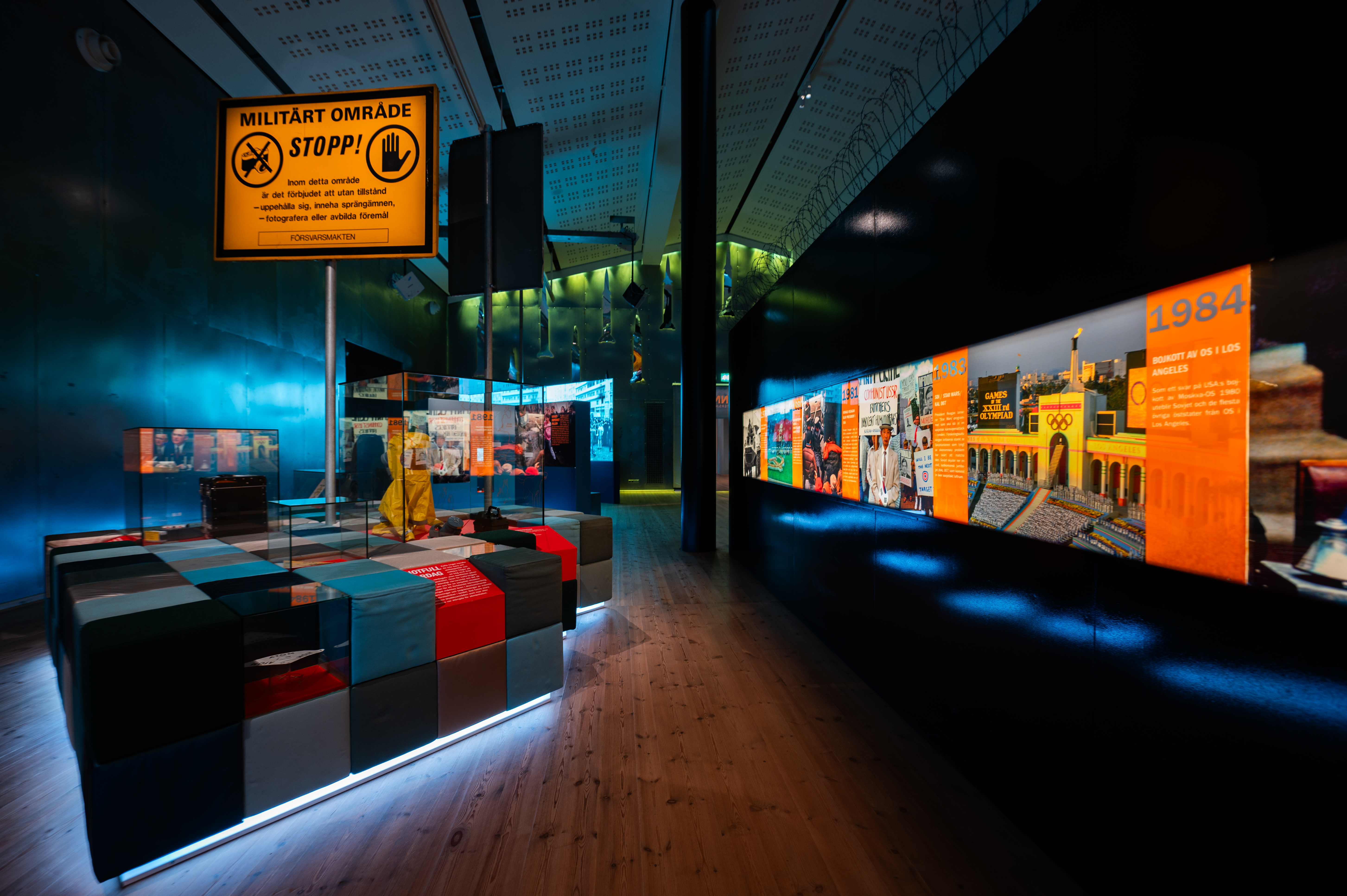 Stort utställningsrum med trägolv och dov belysning. Glasmontrar och orangea skyltar finns i rummet.