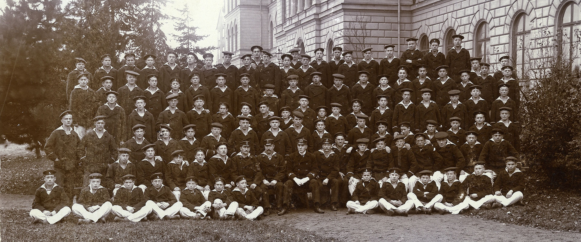 Ett stor grupp pojkar i likadana kläder och mössor. Bakom dem granskog till vänster och en stor stenbyggnad till höger. Gruppbild på skeppsgossekåren.