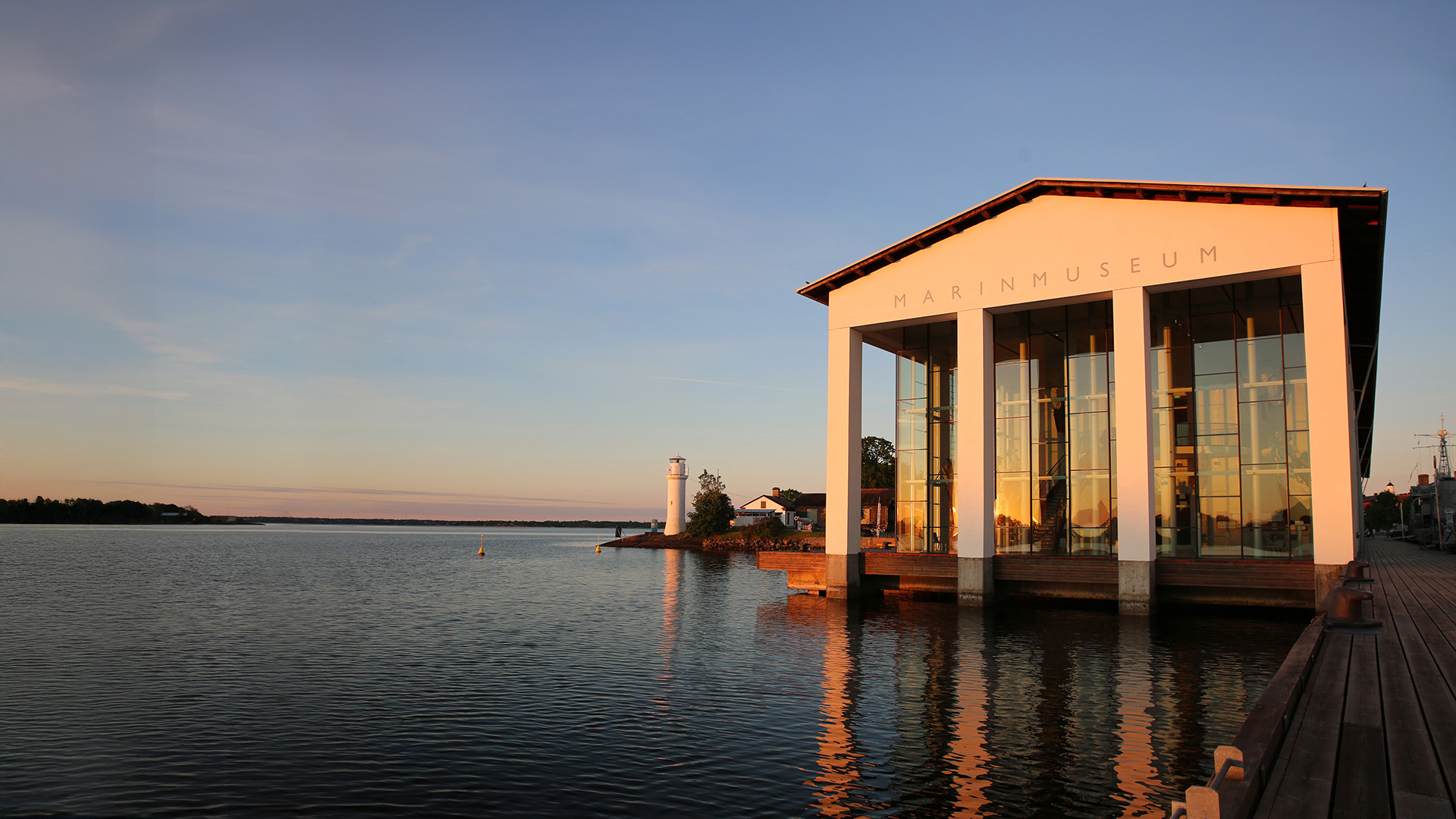 The Naval Museum in Karlskrona by the water at sunset, with warm light reflecting in the large glass windows and a lighthouse in the background.