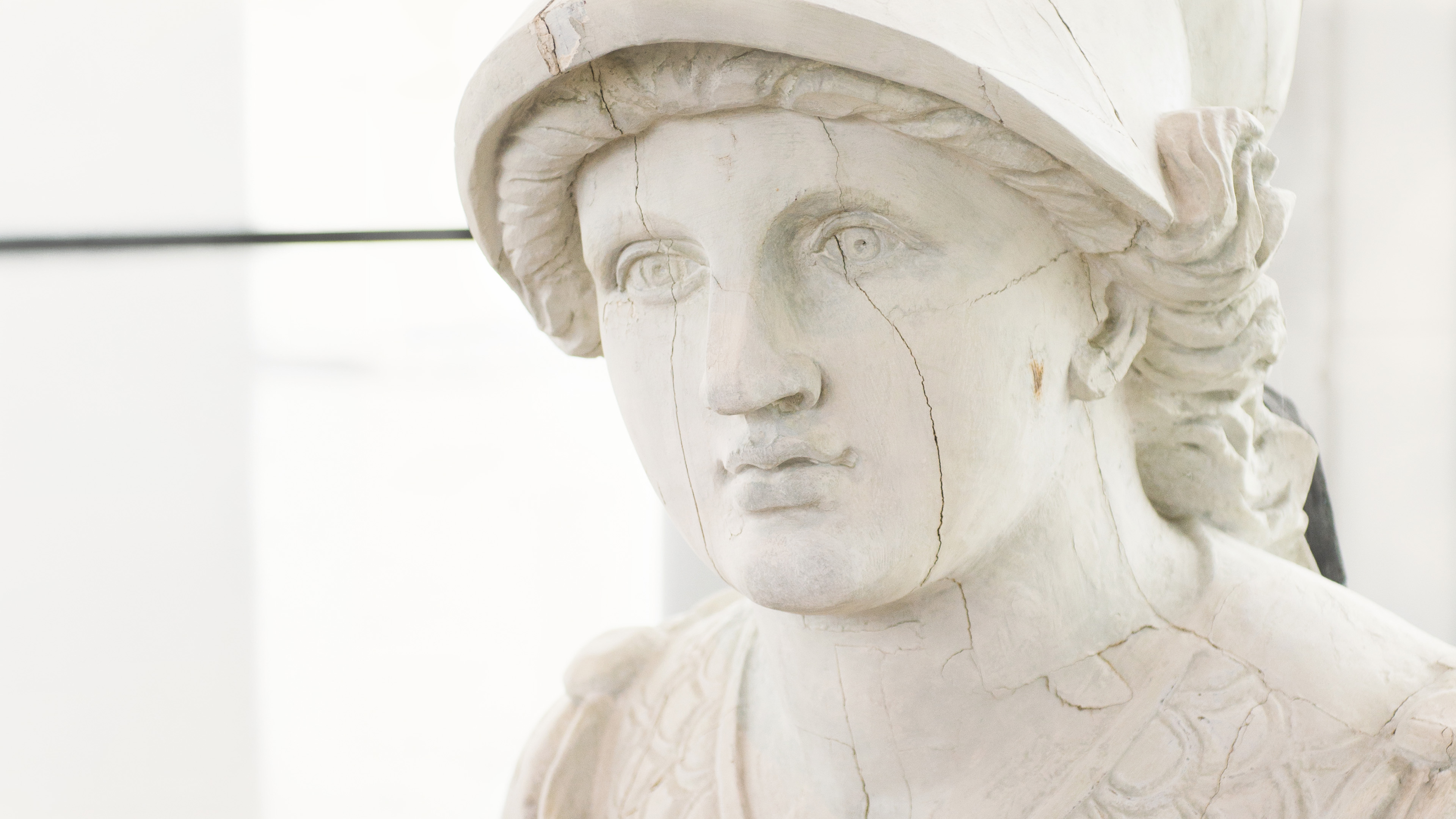 Närbild av en ljus antik skulptur med synliga sprickor och klassiska detaljer, fotograferad i ett ljust inomhusrum.
