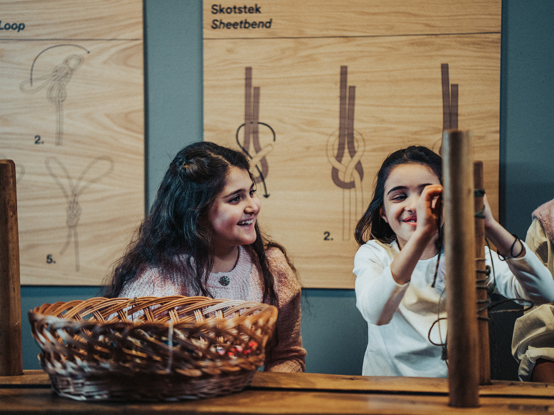 Two children sit at a table in the Naval Museum, learning to tie knots while smiling and concentrating in front of wooden boards showing different knot types.