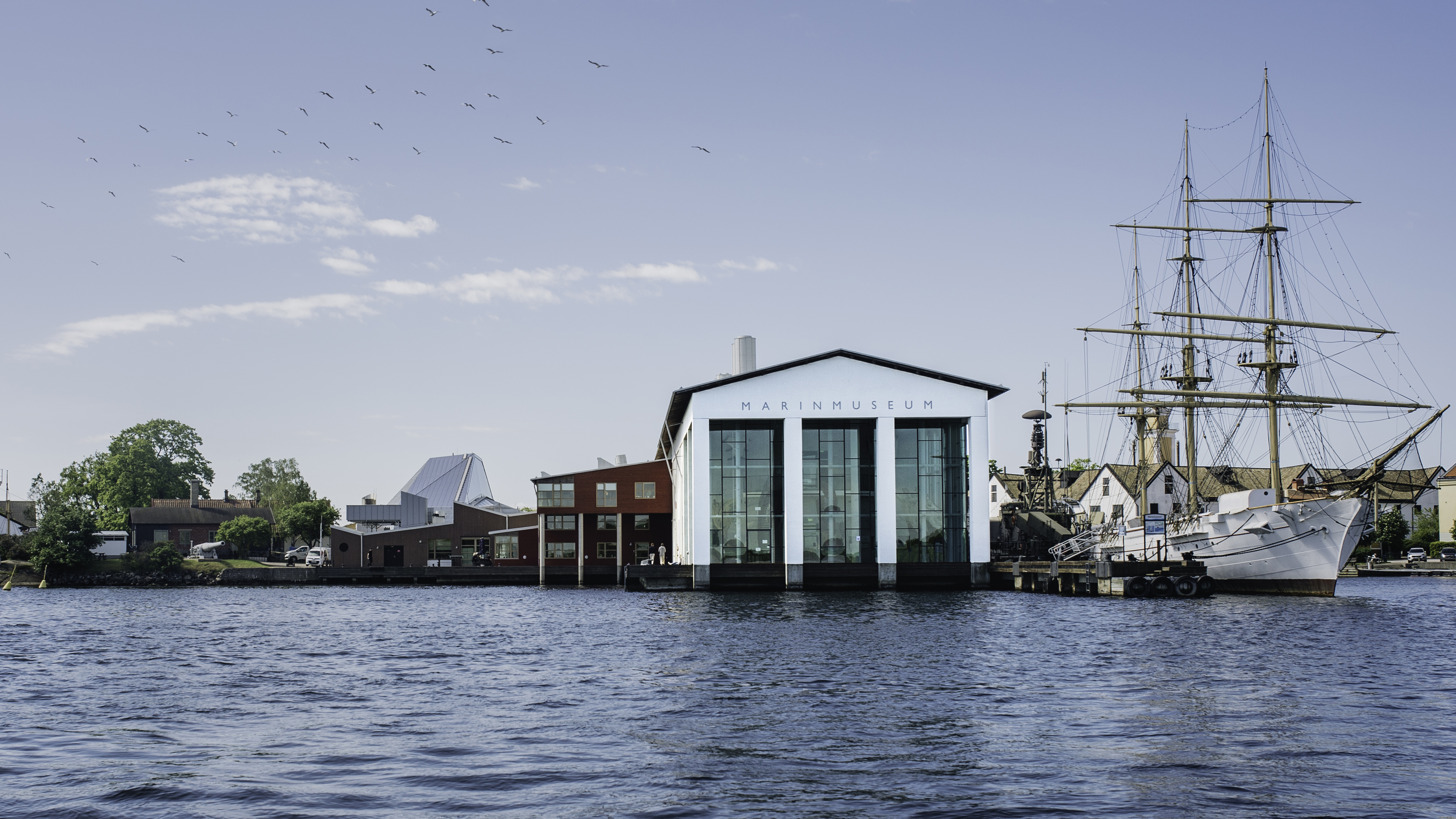 Marinmuseum i Karlskrona sett från vattnet en solig dag, med en segelfartyg förtöjt vid museet och fåglar som flyger på himlen.
