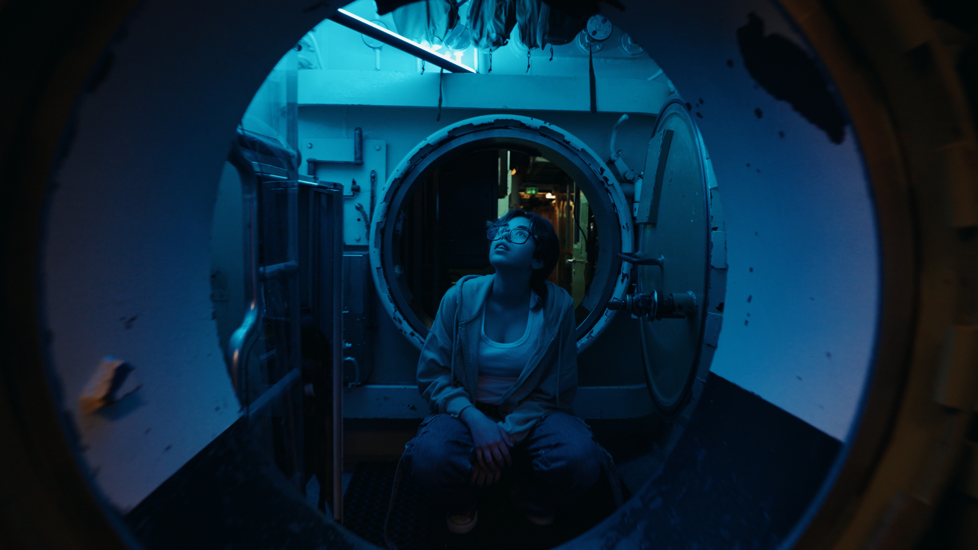 A person wearing glasses sits inside a submarine, illuminated by blue light, looking up at the ceiling through a round metal hatch.