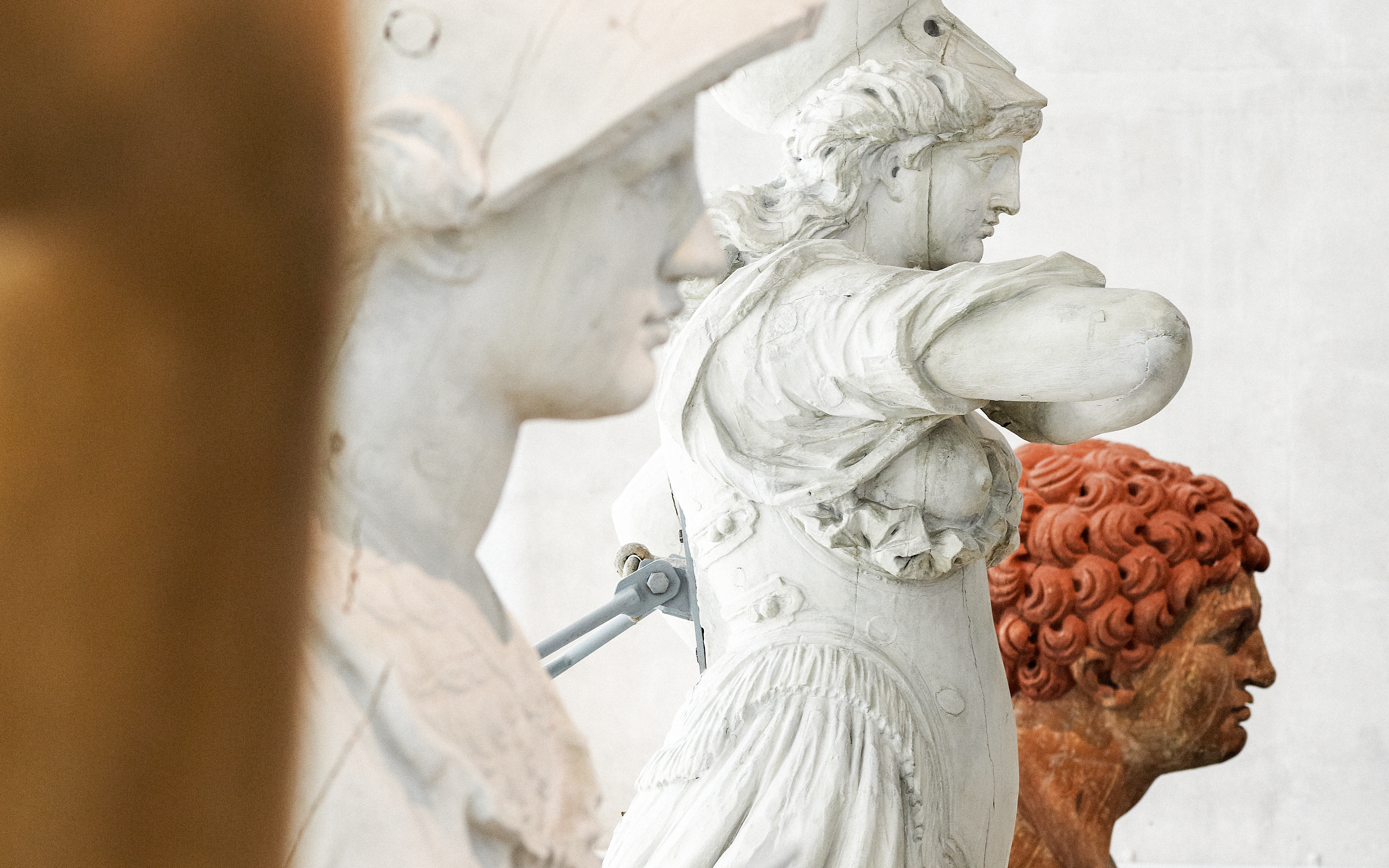 Bright photo of three figureheads seen from the side, the middle one is dressed as a Roman warrior. Two of the figures are white, the third is rust red.